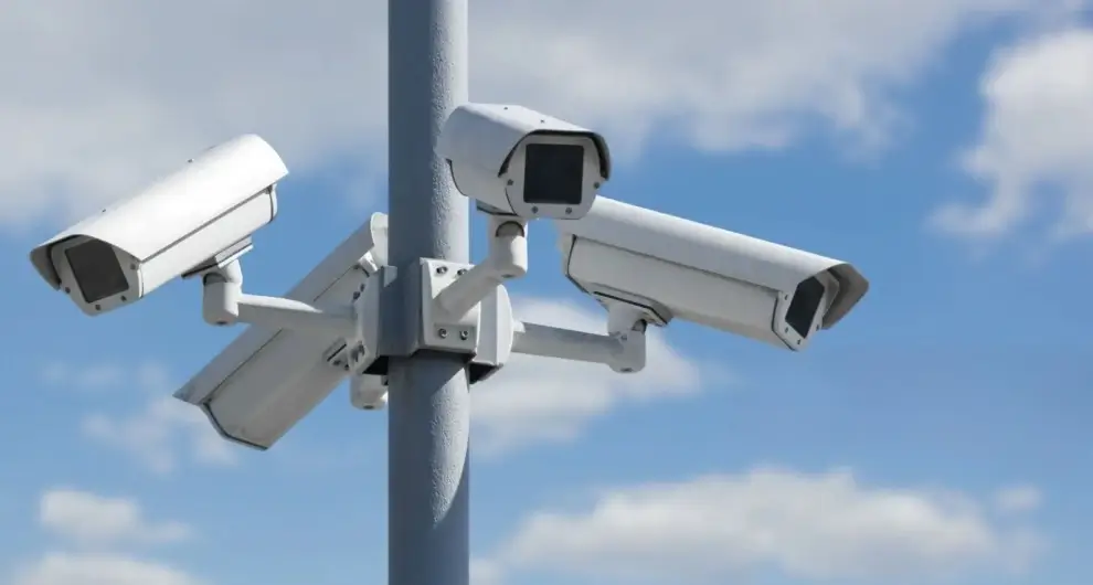 Cluster of four outdoor surveillance cameras mounted on a pole against a blue sky with scattered clouds.