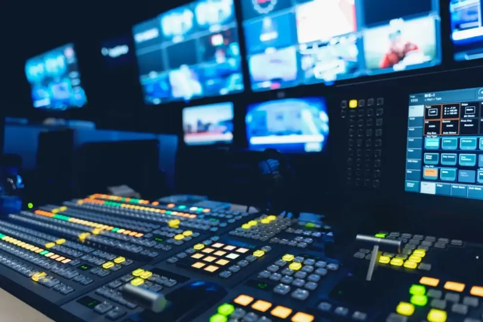 Broadcast control room with illuminated switchboards, monitors displaying live video feeds, and various control panels used for video production and live streaming.