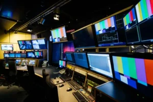 Modern television production control room with multiple monitors displaying color bars, camera feeds, and waveform data, surrounded by workstations equipped with editing and communication equipment.