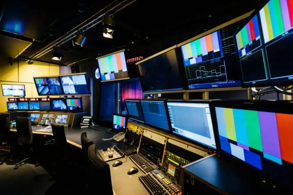 Modern television production control room with multiple monitors displaying color bars, camera feeds, and waveform data, surrounded by workstations equipped with editing and communication equipment.